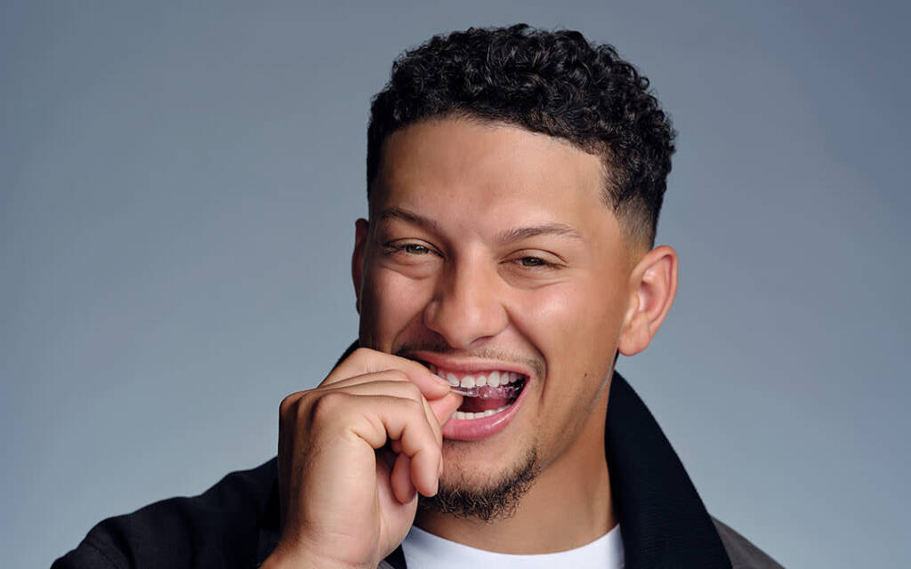 Patrick Mahomes smiling while holding an Invisalign aligner in his mouth against a grey background.